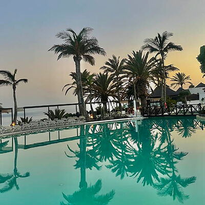 Abendstimmung, Sicht über einen grünlich schimmernden Pool. Im Hintergrund Palmen und das Meer